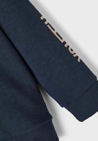 Felpa blu navy con polsini e orlo a costine, con testo color beige chiaro stampato lungo la manica, realizzata in morbido cotone.