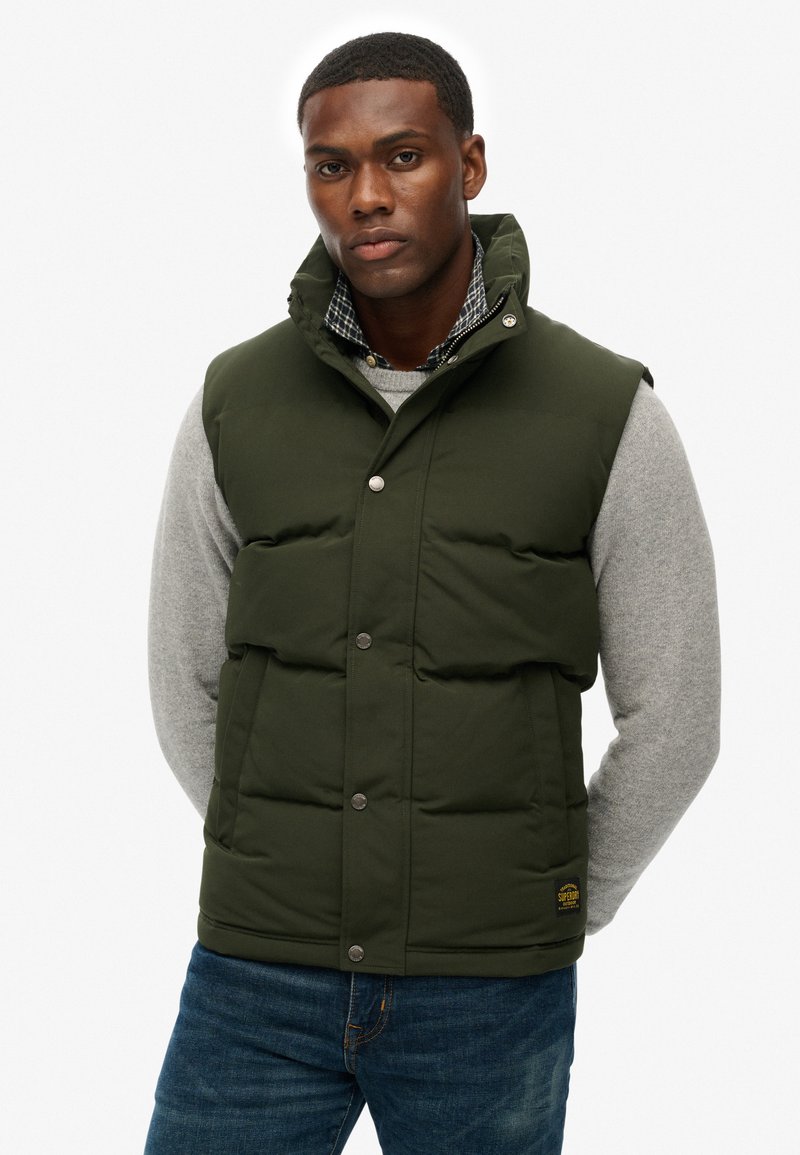 Man die een donkergroene gewatteerde bodywarmer draagt over een grijze trui en een blauwe spijkerbroek, staand tegen een effen witte achtergrond.