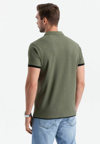 Olivgrön pikétröja med svart kant, kortärmad och klassisk krage, i kombination med ljusblå jeans. Slätt, bomullstyg.