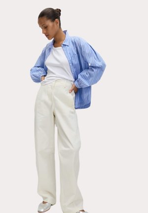 Camicia oversize a righe blu e bianche, t-shirt bianca e pantaloni larghi crema chiaro. Scarpe slip-on argentate. Vestibilità semplice e rilassata.