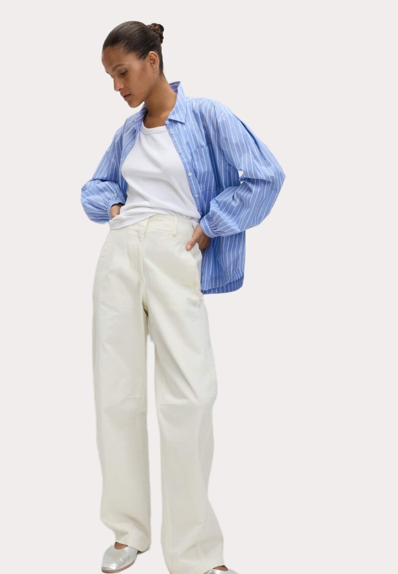 Gestreiftes Übergrößenhemd in Blau und Weiß, weißes T-Shirt und hellcremefarbene, weite Hose. Silbernene Slipper. Einfacher, lockerer Schnitt.