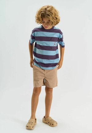 Barn med krøllet blondt hår iført en blå- og lilla stribet skjorte, beige shorts og brune slip-on sko, står og kigger ned.