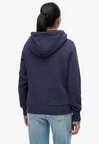 Vrouw met donker haar in een paardenstaart, gekleed in een marineblauwe hoodie en lichtblauwe spijkerbroek, staand met haar rug naar de camera tegen een effen witte achtergrond.