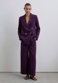 Blazer morado de doble botonadura con botones dorados, combinado con pantalones de pierna ancha en el mismo tejido, que presentan una textura suave y líneas limpias.