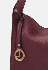 Sac à main en cuir bourgogne foncé avec une texture granuleuse, un accent en métal doré et un charm en forme de logo circulaire distinctif attaché à la sangle.