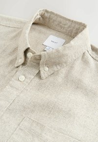 Chemise boutonnée texturée beige clair avec col, comprenant deux boutons sur le devant et une poche poitrine. L'étiquette de la marque indique "NEXT".