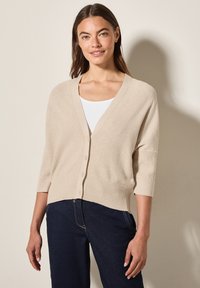 Beige gestrickte Strickjacke mit tiefem V-Ausschnitt, sieben Knöpfen und dreiviertel Ärmeln. Übert einem weißen Oberteil getragen und mit dunklen Jeans kombiniert.