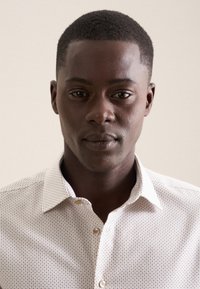 Chemise à boutonnage avec une base beige clair et de petits pois bleus. Présente un col et une texture lisse, vue portée par une personne.