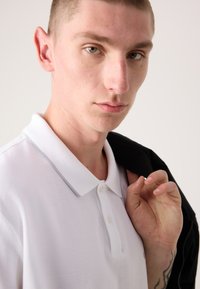 Hvit polo skjorte med teksturert stoff, en krage med grå kant og tre knapper. Modellen holder en svart jakke over den ene skuldren.