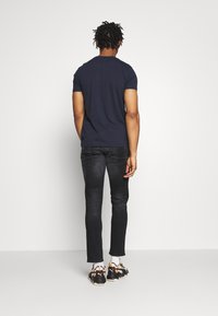 Marinblå kortärmad t-shirt, svarta slim-fit jeans och svarta sneakers med orangea accenter. Bakvy, enfärgad vit bakgrund.