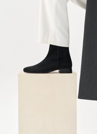 Černé semišové kotníkové boty se špičatou špičkou a nízkým blokovým podpatkem, umístěné na dřevěném podstavci, doplněné o bílé kalhoty.