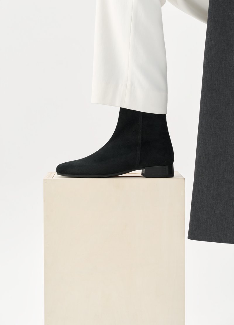 Schwarze Wildleder-Stiefeletten mit spitzem Zehenbereich und niedrigem Blockabsatz, auf einem Holzpodest platziert, kombiniert mit weißen Hosen.