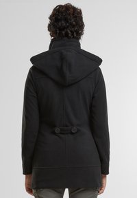 Črni plašč s kapuco iz mehke tkanine. Ima prilegajoč se kroj, velik ovratnik in dva gumbka na zadnjem pasu.