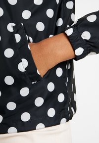 Černá bunda s bílými puntíky, s elastickými manžetami a bočními kapsami, vyrobená z hladkého, lesklého materiálu s na míru střiženým vzhledem.