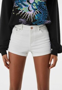 Shorts de mezclilla blanca con cintura alta, puños enrollados y bolsillos frontales. Combinados con una blusa negra de manga larga que presenta un diseño gráfico colorido.