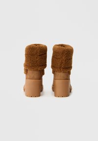 Boots negre din suede cu toc gros și manșete din blană plisată, văzute din spate pe un fundal alb simplu.
