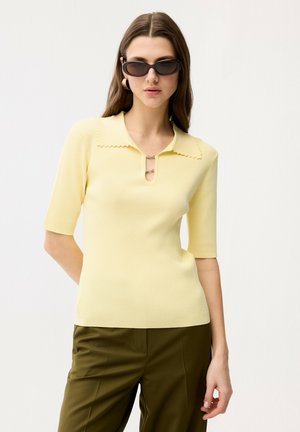 Kvinde iført en lysegul kortærmet top med bølget krave og olivengrønne bukser, accessoriseret med mørke solbriller og øreringe i hoop-stil.