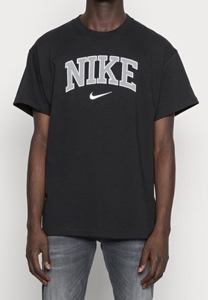 Black short-sleeve T-shirt with large gray "NIKE" text and white swoosh logo worn by person in gray jeans, standing against plain background.
