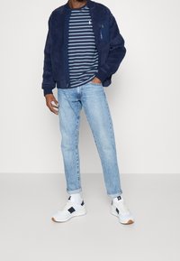 Navy fleece bomber jacket, striped navy and blue T-shirt, light denim jeans, and white sneakers with navy accents. Casual outfit displayed.