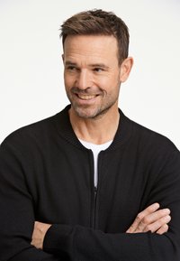 Mustan neulotun kardiganin, jossa on uritettu kaulus ja hihansuut, vetoketjukiinnitys, käytetään valkoisen paidan päällä; siinä on rento istuvuus ja teksturoitu kangas.