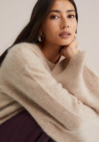 Gai smilškrāsas džemperis mīkstā, pūkainā materiālā ar ribotām piedurknēm. Modelim ir lieli zeltīti gredzena formas auskari un viņš atbalsta zodu uz rokas. Tumši purpura apakša.