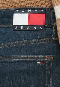 Kék farmer anyag narancssárga varrással; fekete címkével, amelyen a "TOMMY JEANS" felirat és egy téglalap alakú piros-fehér logó látható. Kis logó részlet a zseben.