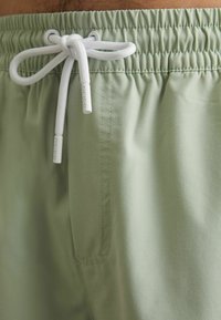 Shorts vert clair avec une taille élastique, cordon de serrage blanc et poche latérale. Texture lisse avec des détails de couture soignés.