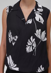 Blusa senza maniche nera con un motivo floreale bianco, dotata di un collo alla coreana e di un tessuto morbido e liscio. Design drappeggiato sulla parte anteriore.