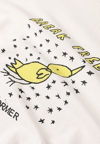 Fehér pamut póló, sárga madár illusztrációval, fekete csillagokkal körülvéve, a "BREAK FREE" felirattal fent és a "FARMER" felirattal lent.