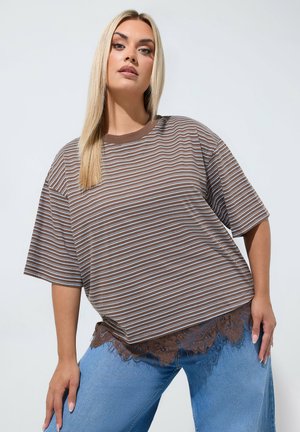 Jonge vrouw met lang blond haar draagt een bruin gestreept los t-shirt over een blauwe jeans met bruine kanten rand tegen een effen lichte achtergrond.