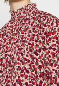 Blommig blus med hög smockad krage, tillverkad av lätt tyg med rosa bakgrund och flerfärgade blommönster i rött och grönt.