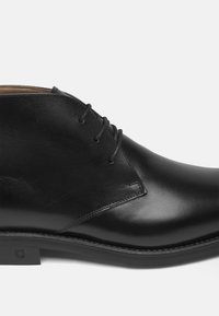 Scarpa in pelle nera con una finitura liscia, caratterizzata da un profilo basso, punta tonda e tre occhielli per i lacci. Cuciture decorative lungo la suola.