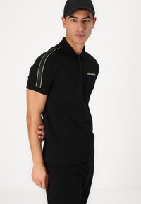 Svart polo t-shirt i bomull, med krage, korta ärmar, guldlogotyp och kontrasterande gröna sidränder.