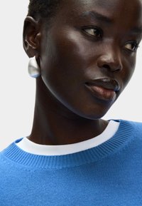 Modrý pletený svetr s kulatým výstřihem na bílé vrstvě, doplněný bílou kapkovou náušnicí, vytvářející hladký, moderní vzhled.