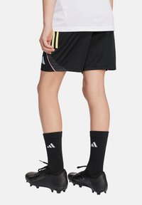 Schwarze Sportshorts mit gelben Streifen, kombiniert mit schwarzen Socken und Fußballschuhen. Das Design zeigt geschwungene Akzente und ein Logo auf den Shorts.