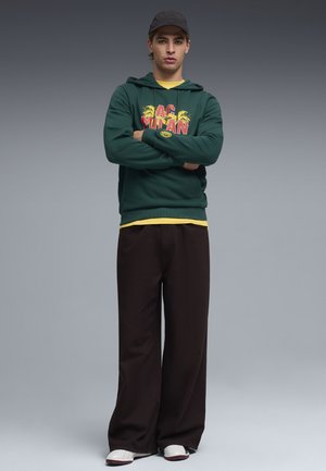 Felpa verde con grafica rossa e dettagli gialli, abbinata a pantaloni neri a gamba larga e sandali bianchi. Tessuto morbido con polsini a coste.