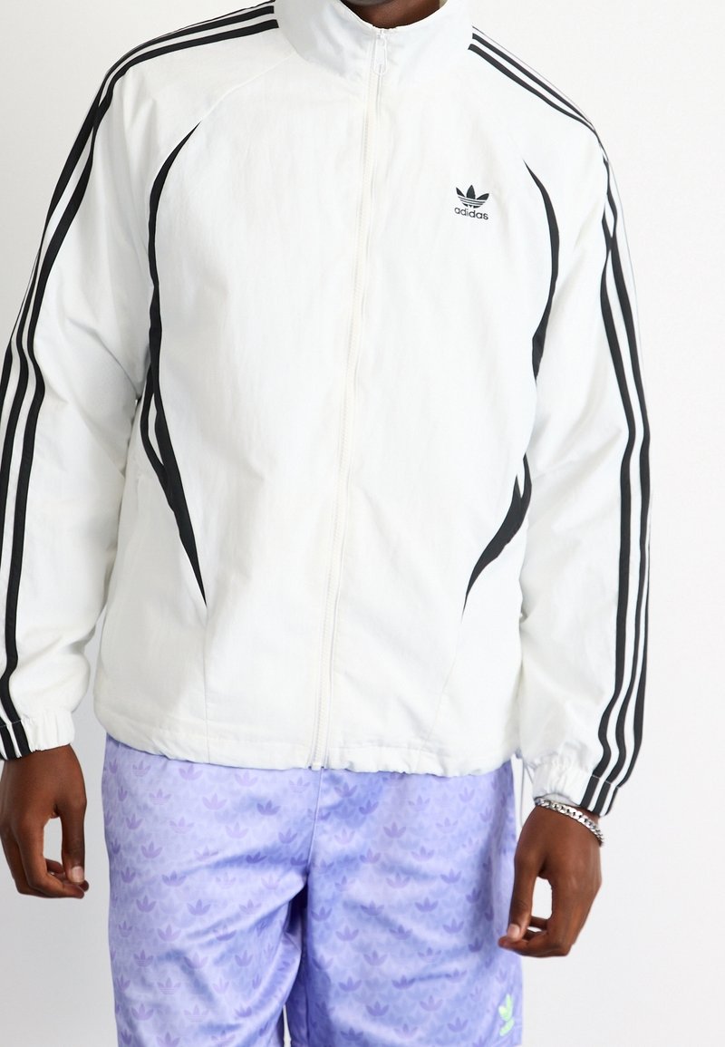Vit zip-up jacka med svarta ränder längs axlarna och armarna, med en Adidas-logotyp på vänster bröst. Texturerat tyg.