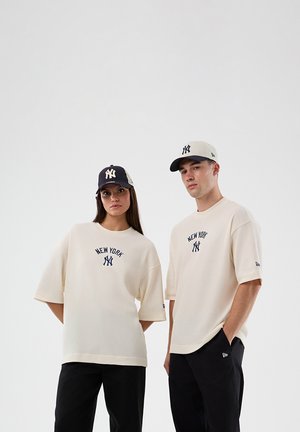 Dvoje ljudi u bež majicama New York Yankees i ujednačenim bejzbol kapama stoje na jednostavnoj svijetloj pozadini.