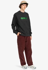 Black sweatshirt with green graphic logo, paired with burgundy corduroy pants, and gray sneakers. Casual outfit with contrasting colors.