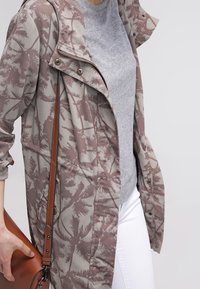 Lätt jacka i mönstrad mauve och beige tyg, med dragkedja fram, huva och knappslå, tillsammans med en grå topp och vita byxor.