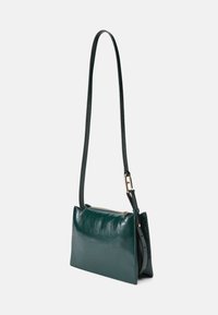 Grön läder crossbody-väska med en mjuk textur, smal rektangulär form, justerbar axelrem och en dragkedja i guldton.