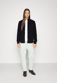 Zwarte button-up shirt met een rood logo, gedragen over een wit T-shirt, gecombineerd met lichte spijkerbroeken en zwarte schoenen. Soepele textuur, casual pasvorm.