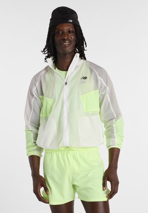 Mann mit dunkler Haut und Dreadlocks, der eine schwarze Mütze, eine transparente weiße Jacke, ein neongrünes Hemd und passende neongrüne Shorts trägt.