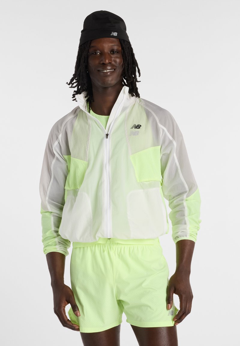 Homme à la peau foncée et aux dreadlocks, portant une casquette noire, une veste blanche translucide, un t-shirt vert néon et un short vert néon assorti.