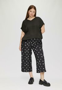 Schwarzes, weit geschnittenes Oberteil mit V-Ausschnitt, kombiniert mit schwarzen, gemusterten Culottes, die weiße abstrakte Formen aufweisen. Schwarze, klobige Loafers runden den Look ab.