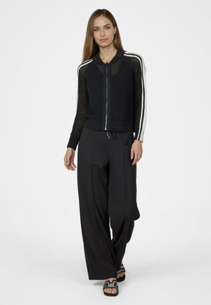 Woman wearing a black mesh zip-up jacket with white stripes on sleeves, wide-leg black pants, and black sandals with decorative straps.