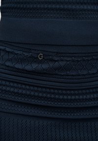 Gros plan sur un tissu tricoté bleu foncé avec des textures variées, comprenant des côtes, un mesh, un motif en losange, et un petit détail métallique en forme de "G".