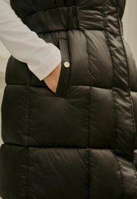 Vest cu umplutură neagră, cu un design cu curea, buzunare laterale cu accente din butoane și textură matlasată. Cămașă albă cu mânecă lungă vizibilă dedesubt.