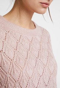 Maglione lavorato a maglia rosa chiaro con un motivo a rombi e scollatura rotonda. La texture è morbida e dettagliata con punti aperti.
