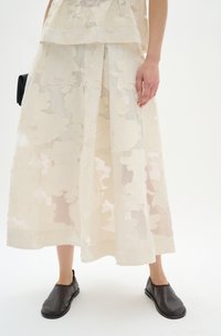 Ein transparenter, geschichteter Maxirock in Elfenbein mit strukturierten Blumenmustern, kombiniert mit schwarzen Slippern aus Leder und einer kleinen schwarzen Tasche.